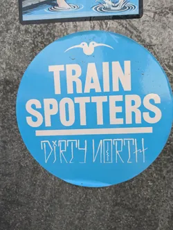 Street sticker A round blue sticker with the text "TRAIN SPOTTERS" in white uppercase letters. Underneath, "DIRTY NORTH" is written in a stylized font.  A small bird-like symbol is at the top.