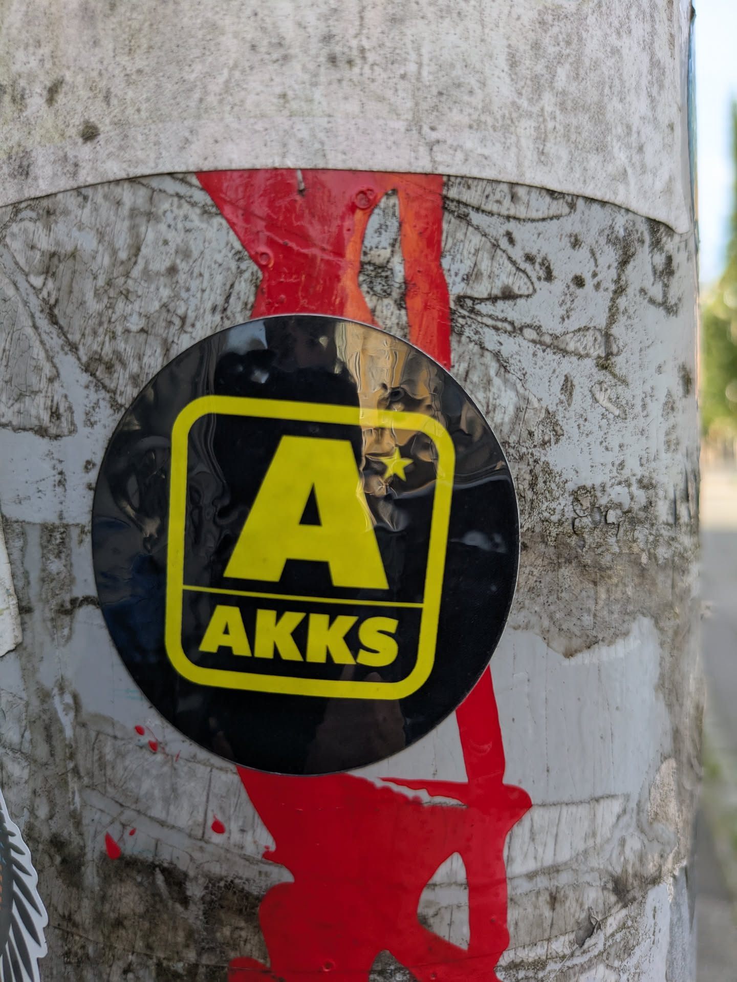 The image features a black circular sticker adhered to a weathered, light-colored pole. The sticker has a prominent yellow 'A' enclosed within a rounded square frame, with a small yellow star integrated into the top right corner of the 'A'. Below the 'A', the text 'AKKS' is written in yellow uppercase letters. The pole itself shows signs of wear, with dirt, grime, and patches of what appears to be dried red paint or residue, suggesting previous layers of paint or other markings. There are also visible textures and potential remnants of other old stickers or paper on the pole.