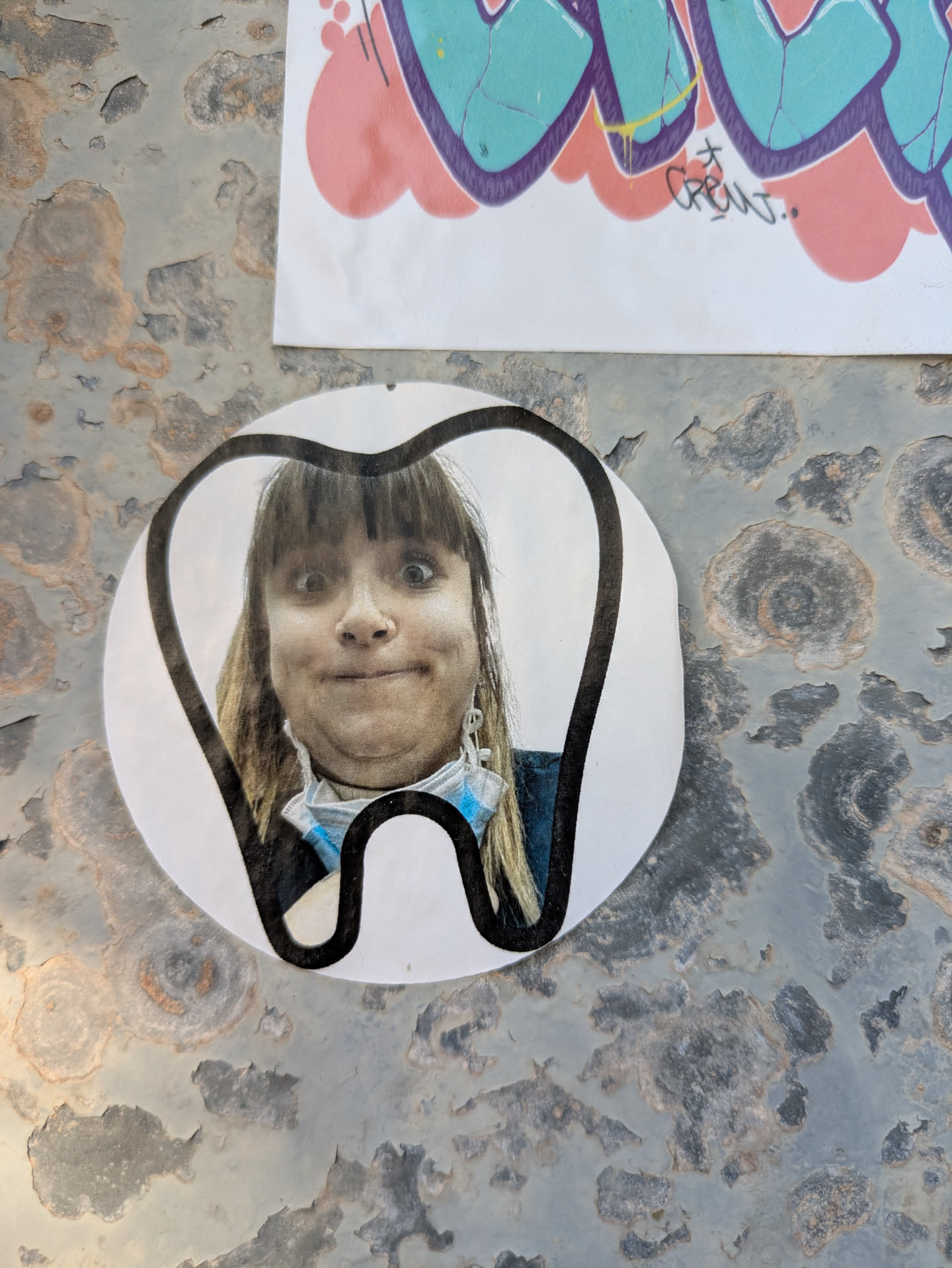 A circular sticker depicting a tooth shape, with the face of a person making a surprised or silly face inside. The person has light skin, bangs, and long hair, and is wearing a mask around their neck and diamond shaped earrings. The sticker is adhered to a rusty metal surface.