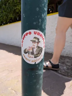 Street sticker An oval white sticker on a green metal post. It features a central illustration of a mustachioed man in a Tyrolean hat holding a beer mug. Red text arches over the top, and gold text arches along the bottom.