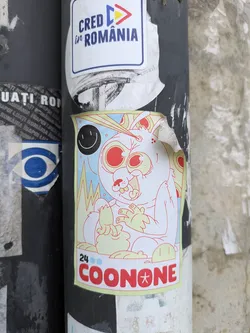 Street sticker This sticker depicts a cartoonish, rabbit-like creature with large, red eyes and a somewhat unsettling expression. It's holding something in its hands. The sticker also features a small, simple smiley face and the text "24 COONONE", with the "O" in "COONONE" replaced by a star. The style is reminiscent of underground or independent comics.