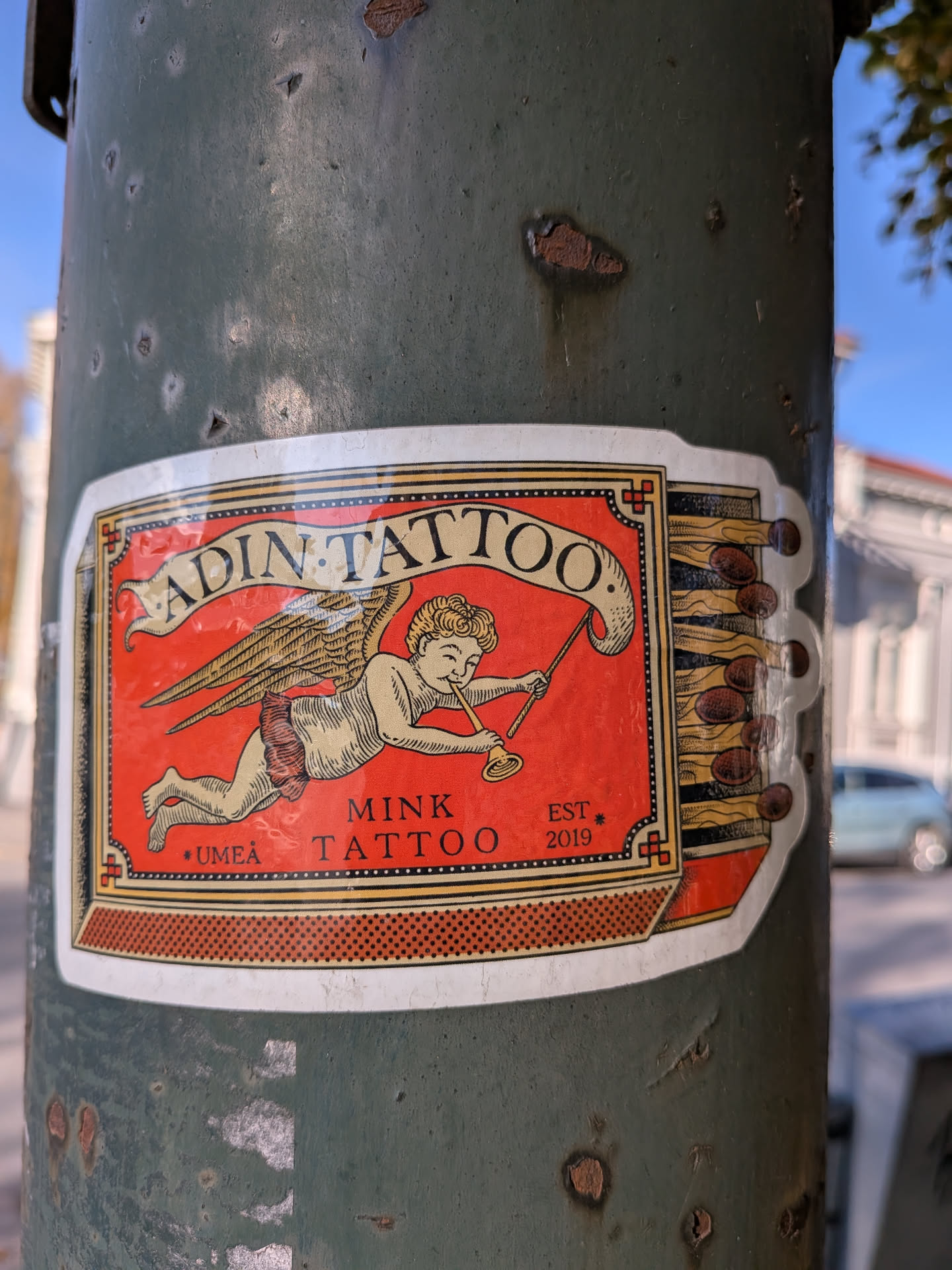 The sticker is predominantly red with a white border. At its center is an illustration of a winged cherub with blonde curly hair, wearing a red loincloth, playing a golden trumpet. The cherub is depicted in mid-flight. Above the cherub, a banner unfurls with the text 'ADIN TATTOO' in black, stylized font. Below the cherub, the words 'MINK TATTOO' are written in black, with '* UMEÅ' to the left and 'EST * 2019' to the right. The right side of the sticker features an illustration of a row of unlit matches with brown heads and yellow sticks. The overall design has a vintage, illustrative aesthetic.