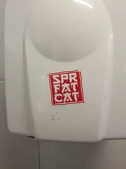 Street sticker A square sticker with a red background and white border, featuring the vertically stacked text 'SPR FAT CAT' in a dripping, graffiti-style font. It is affixed to a white plastic surface and shows slight edge wear.