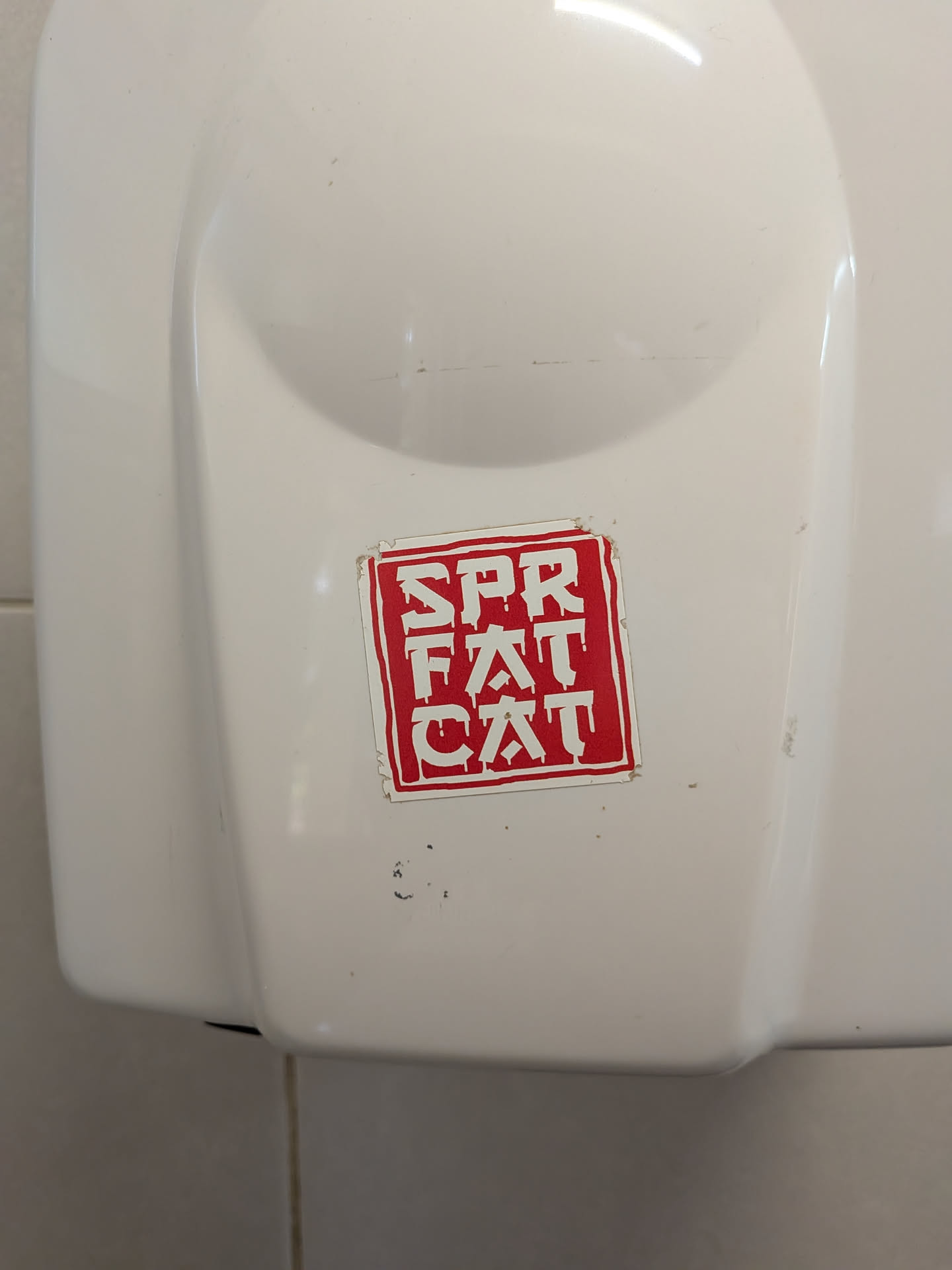 A square sticker with a red background and white border, featuring the vertically stacked text 'SPR FAT CAT' in a dripping, graffiti-style font. It is affixed to a white plastic surface and shows slight edge wear.