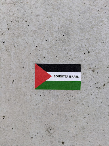 A sticker on a gray concrete surface. The sticker is in the shape of the Palestinian flag. It features a triangle of red color and a stripe of white between black and green stripes. The words "BOJKOTTA ISRAEL" are printed over the white stripe.