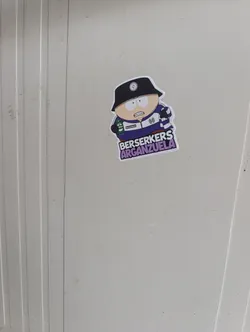Street sticker The sticker features a cartoon character strongly resembling Eric Cartman from South Park, depicted with light skin, wide white eyes, and an angry or determined expression showing clenched teeth. He is wearing a black bucket hat adorned with a white circular logo (possibly a stylized 'M' or 'W') on the front. His attire includes a purple and white striped track jacket or rugby shirt, featuring a white collar and a similar circular logo on the left chest. In his left hand, he holds a green can, which appears to be a beer can with a red 'M' logo and the word 'berserkers' printed on it. Below the character, in bold, blocky, purple letters with a white outline, are the words 'BERSERKERS' on the top line and 'ARGANZUELA' on the bottom line. The entire sticker has a distinct white border, indicating it is die-cut and applied to a light grey, ribbed surface.