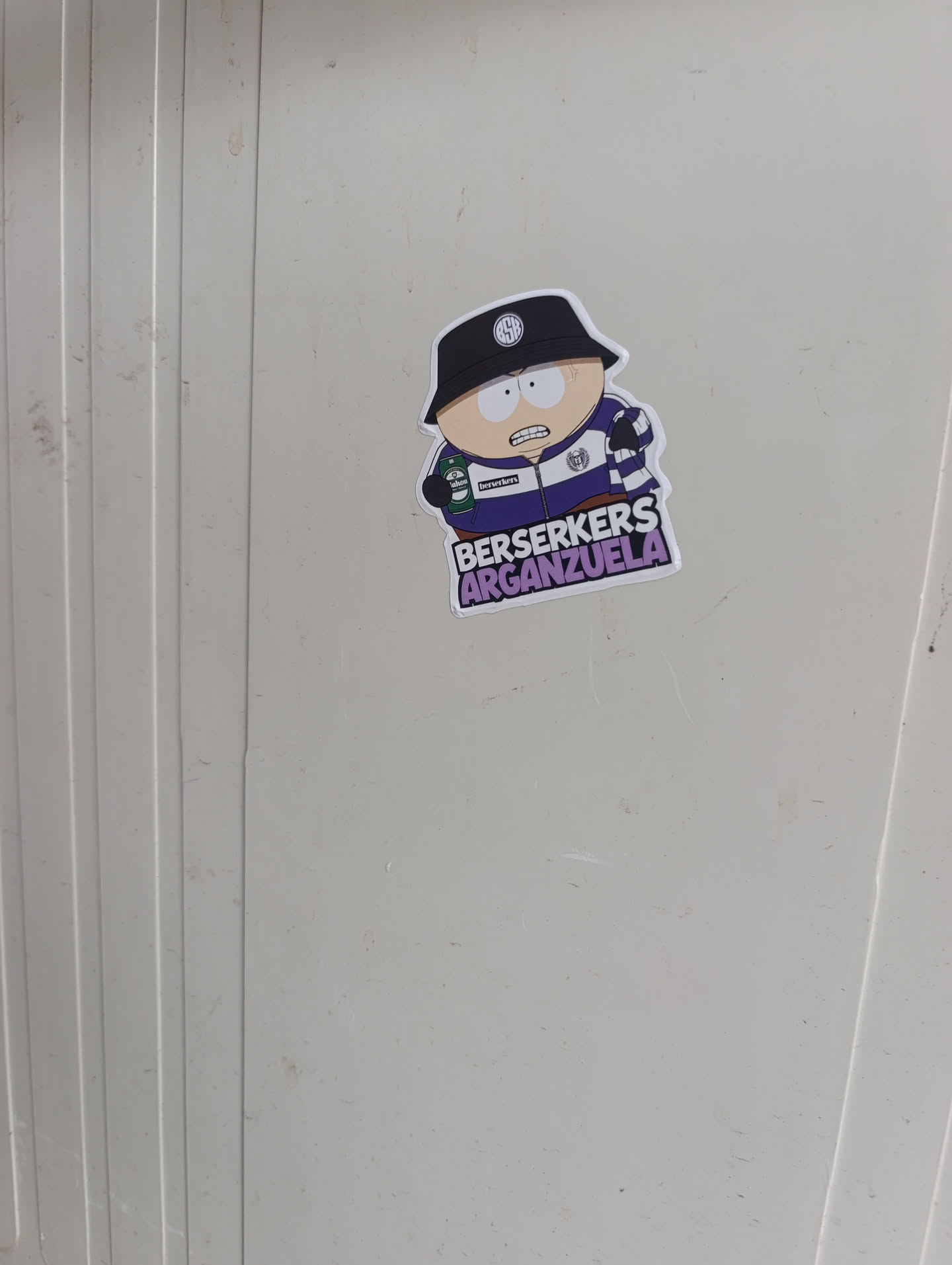 The sticker features a cartoon character strongly resembling Eric Cartman from South Park, depicted with light skin, wide white eyes, and an angry or determined expression showing clenched teeth. He is wearing a black bucket hat adorned with a white circular logo (possibly a stylized 'M' or 'W') on the front. His attire includes a purple and white striped track jacket or rugby shirt, featuring a white collar and a similar circular logo on the left chest. In his left hand, he holds a green can, which appears to be a beer can with a red 'M' logo and the word 'berserkers' printed on it. Below the character, in bold, blocky, purple letters with a white outline, are the words 'BERSERKERS' on the top line and 'ARGANZUELA' on the bottom line. The entire sticker has a distinct white border, indicating it is die-cut and applied to a light grey, ribbed surface.