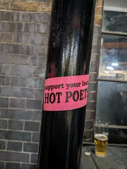 Street sticker The sticker is a rectangular strip with a vibrant, slightly faded pink background. It features bold, black, serif-style text. The top line reads "support your local" in a smaller font, while the bottom line prominently displays "HOT POETS" in a larger, more impactful font. The sticker shows signs of wear, including creases and minor scuffs, indicating it has been affixed to the glossy black pole for some time.