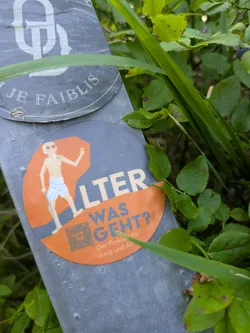 Street sticker The sticker is round with an orange octagonal center and a gray outer ring. It depicts a simplified cartoon of a slender, elderly man with a bald head, sunglasses, and a white loincloth, holding a cane and raising one hand in a wave. The text "ALTER WAS GEHT?" is prominently displayed in a large, bold font on the orange background, with "ALTER" in white and "WAS GEHT?" in a darker gray/blue. Below this, in smaller text, it reads "Der Podcast für Jung und Alt". A QR code is situated to the left of the smaller text.