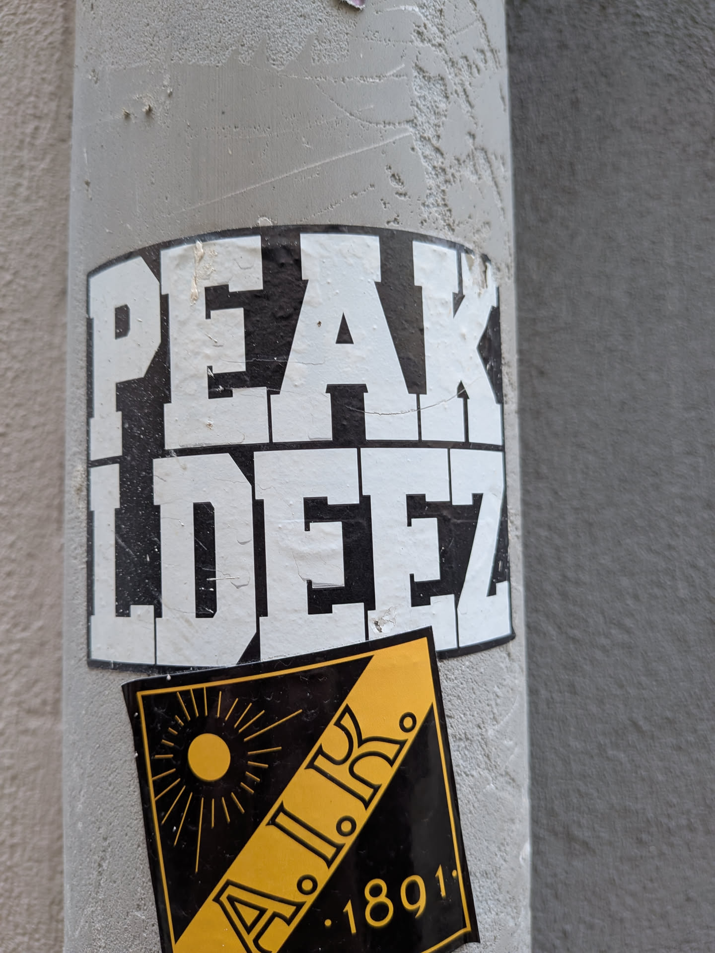 A rectangular black sticker featuring the text 'PEAK LDEEZ' in bold, white block letters. It is stuck to a grey pole and partially overlapped by a yellow and black sticker.