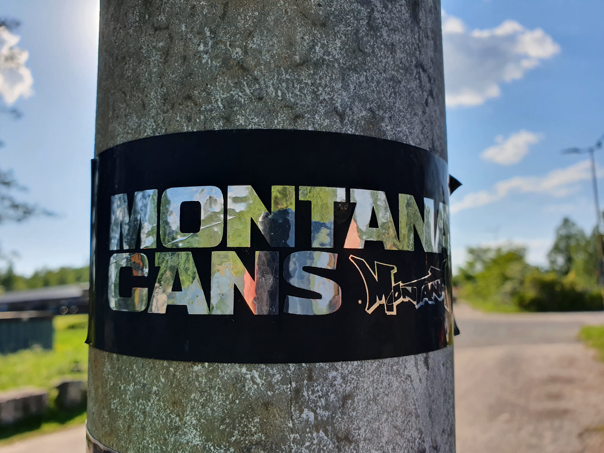 A sticker featuring the Montana Cans logo. The logo is in silver on a black background. The sticker is affixed to a gray pole.
