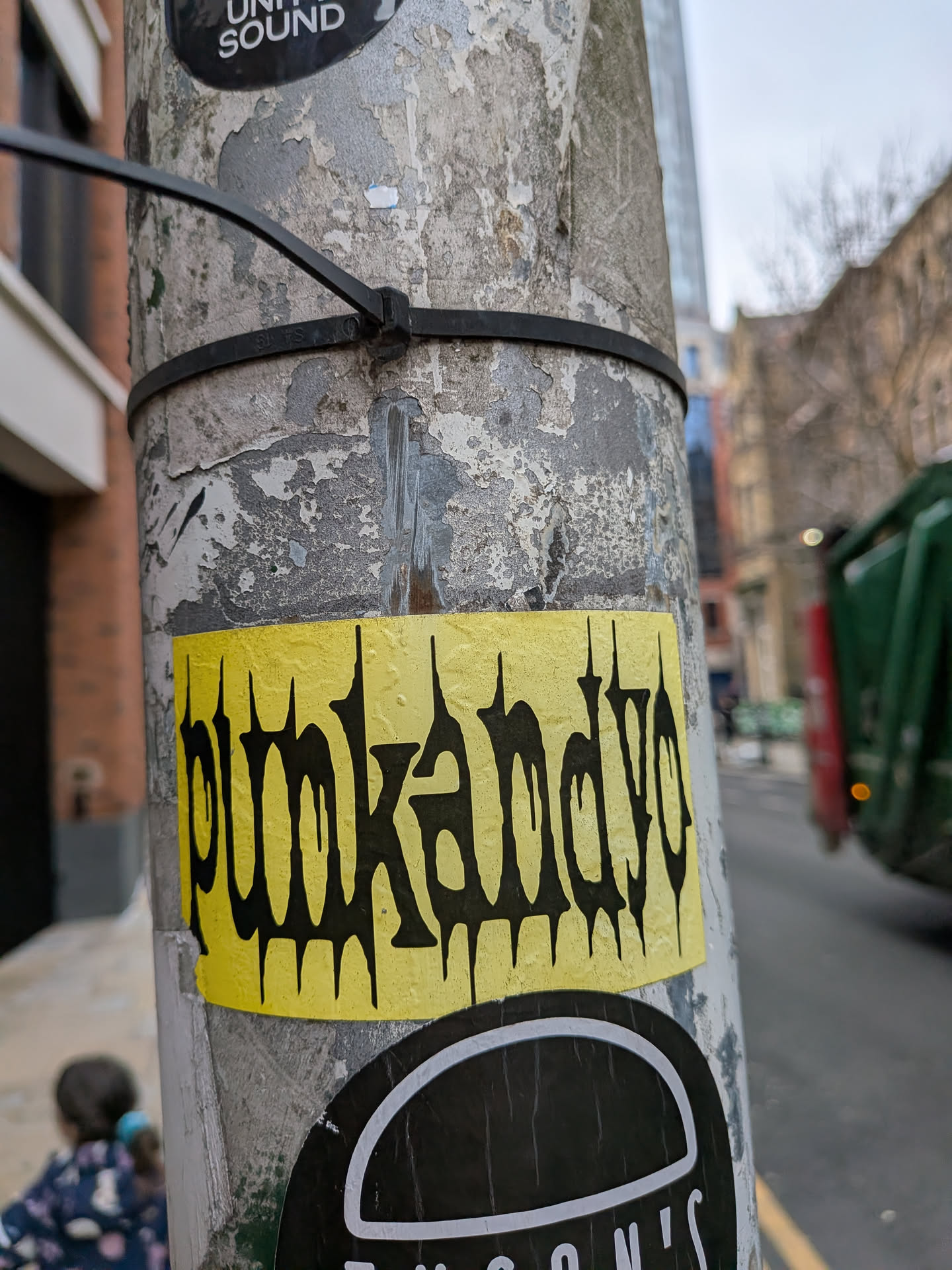 The sticker is a horizontal rectangle with slightly rounded corners, featuring a vibrant, somewhat faded yellow background. Prominently displayed in the center is the word 'punkandyo' in black, stylized text. The font has a distinct jagged, dripping, and somewhat aggressive appearance, reminiscent of punk rock or graffiti aesthetics. The surface of the yellow background shows some texture and minor wear, indicating it's an actual sticker adhered to a weathered pole.