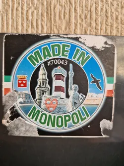 Street sticker A sticker featuring landmarks of Monopoli, Italy. The sticker has a circular design with the text "Made in Monopoli" in green lettering. Inside the circle are images of a lighthouse, a church, and a statue, along with a small red heart. There is a small code #70043 visible on the sticker.