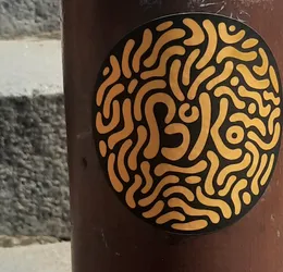 Street sticker The sticker is circular with a solid black background. It features a dense, intricate pattern of flowing, interconnected, organic shapes and lines in a bright, golden-yellow color. The shapes resemble a complex maze or a network of swirling, rounded pathways, filling the entire circular area without any discernible beginning or end. The lines vary in thickness and form, creating a dynamic, almost fluid appearance. The overall impression is abstract and somewhat hypnotic.