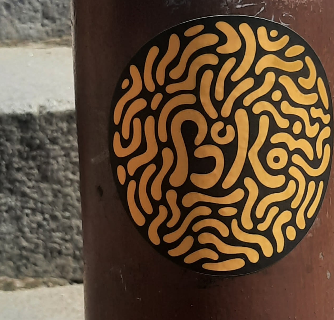 The sticker is circular with a solid black background. It features a dense, intricate pattern of flowing, interconnected, organic shapes and lines in a bright, golden-yellow color. The shapes resemble a complex maze or a network of swirling, rounded pathways, filling the entire circular area without any discernible beginning or end. The lines vary in thickness and form, creating a dynamic, almost fluid appearance. The overall impression is abstract and somewhat hypnotic.
