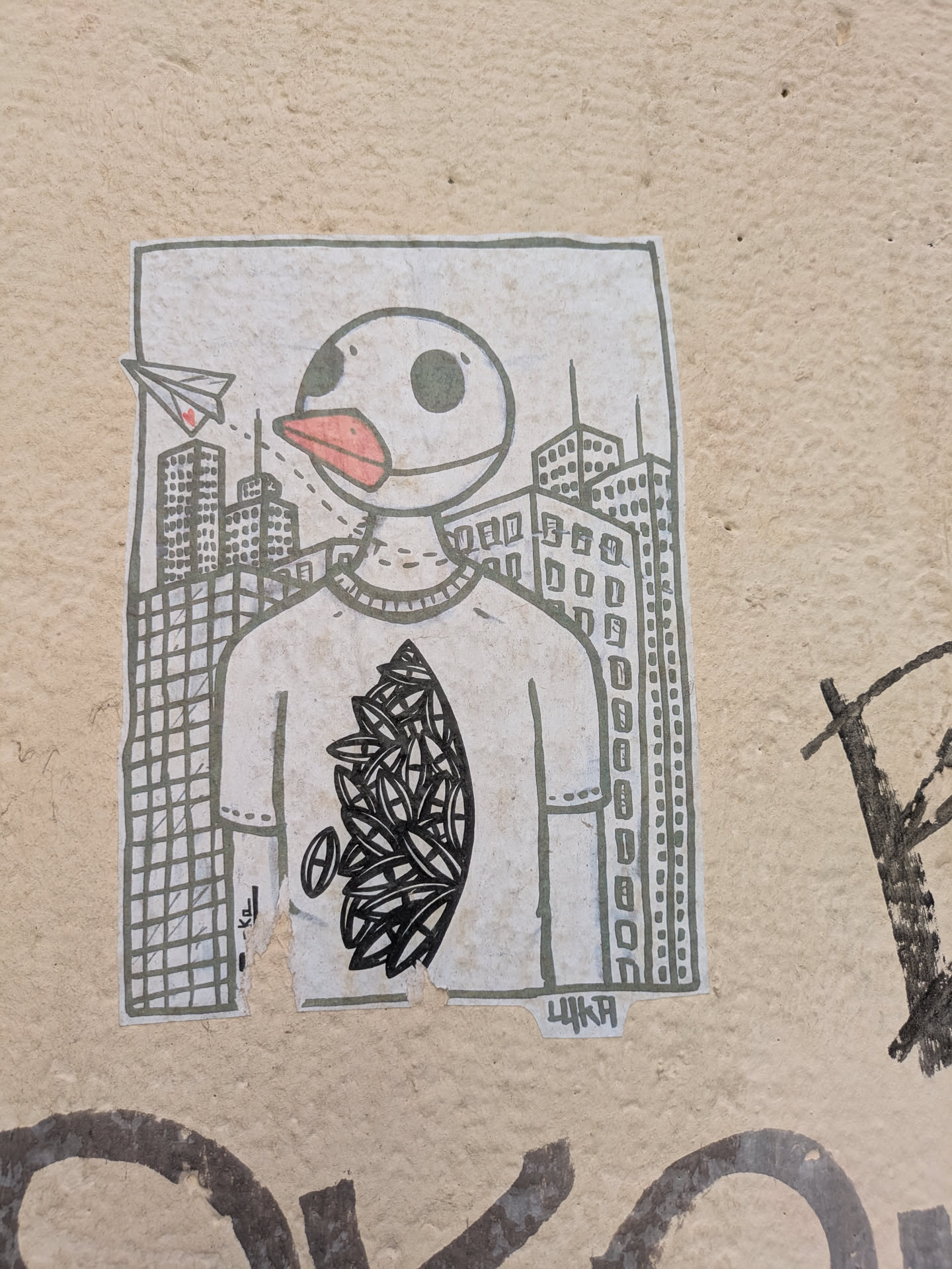 The sticker portrays a bird-headed character wearing a t-shirt, standing against a backdrop of city skyscrapers. A paper airplane with a heart trails behind it, seemingly connecting the bird-like figure to the city. The t-shirt design includes leaf-like shapes. The sticker seems to be hand-drawn in black and white and is damaged on the lower edge.