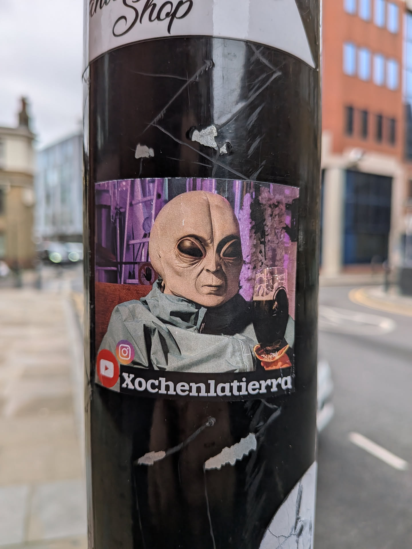 The sticker features a humanoid alien figure with a smooth, light beige head, large dark eyes, and a subtle mouth. It is depicted wearing a grey-green, high-collared jacket. The alien holds a glass of dark beer with a foamy head in its gloved left hand. The background is a blurred, abstract scene dominated by purple hues, suggesting an industrial or futuristic setting. In the bottom left corner, there are visible red YouTube and orange-purple Instagram social media logos. Below the alien's hand, a small, orange, possibly food item or snack is visible. The sticker itself shows minor wear, including slight creases and lifted edges, indicating it's a physical sticker applied to a surface.