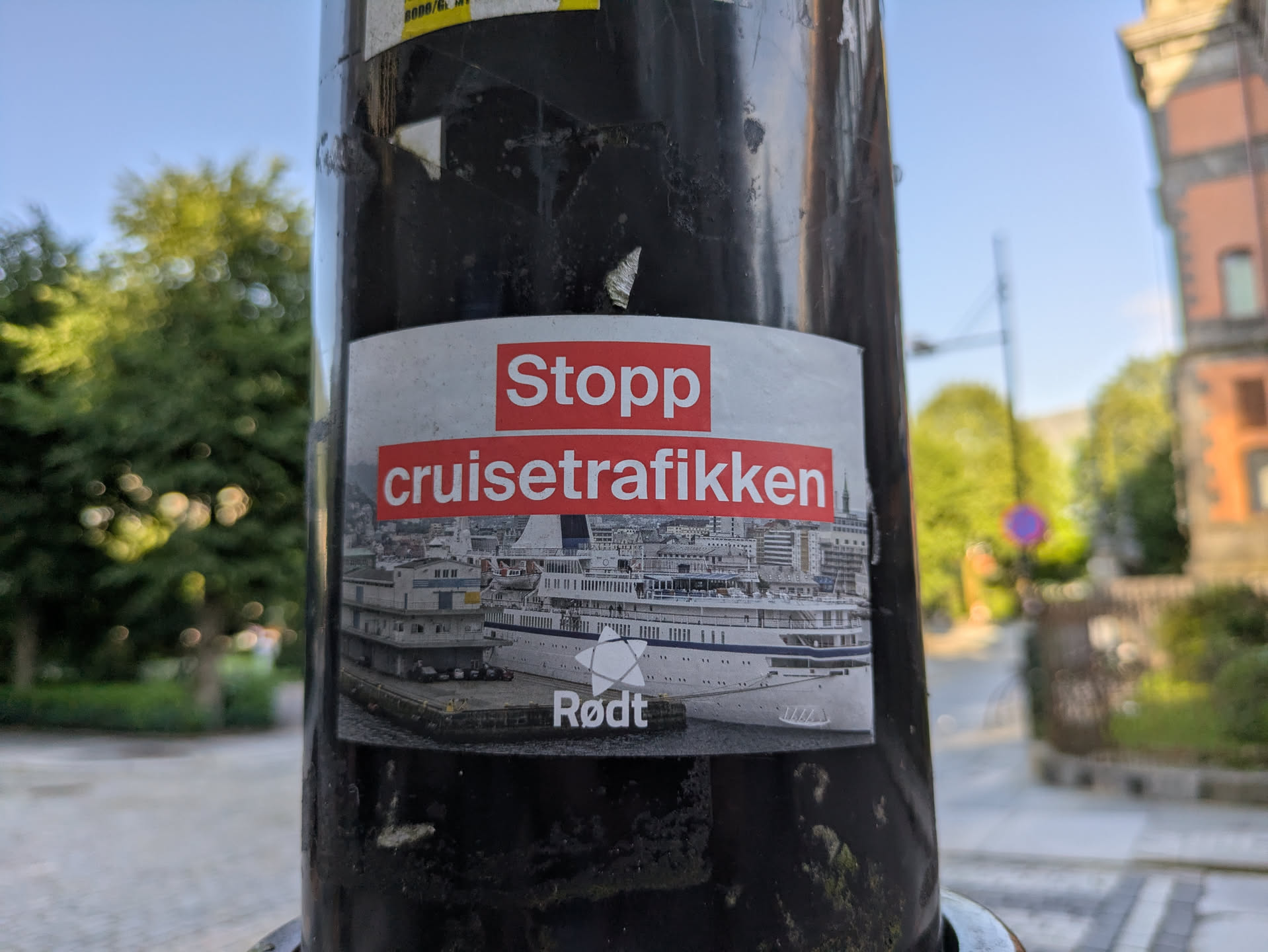 The sticker, placed on a black pole, features a prominent image of a large white cruise ship docked in what appears to be a European city harbor, with buildings and a bustling waterfront visible in the background. Above the image, in white text on a red background, are the words 'Stopp cruisetrafikken'. Below the ship image, also in white text but with a star logo, is 'Rødt'. The sticker is slightly worn at the edges, indicating it has been on the pole for some time. The background outside the pole shows trees, sky, and hints of other urban structures.