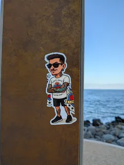Street sticker A die-cut sticker featuring a cartoon caricature of a man with spiky hair, sunglasses, and crossed arms. He is heavily tattooed on his arms and legs, wearing a white 'LOVE ARTE TATTOO-SHOP' t-shirt, black shorts, and black sneakers.