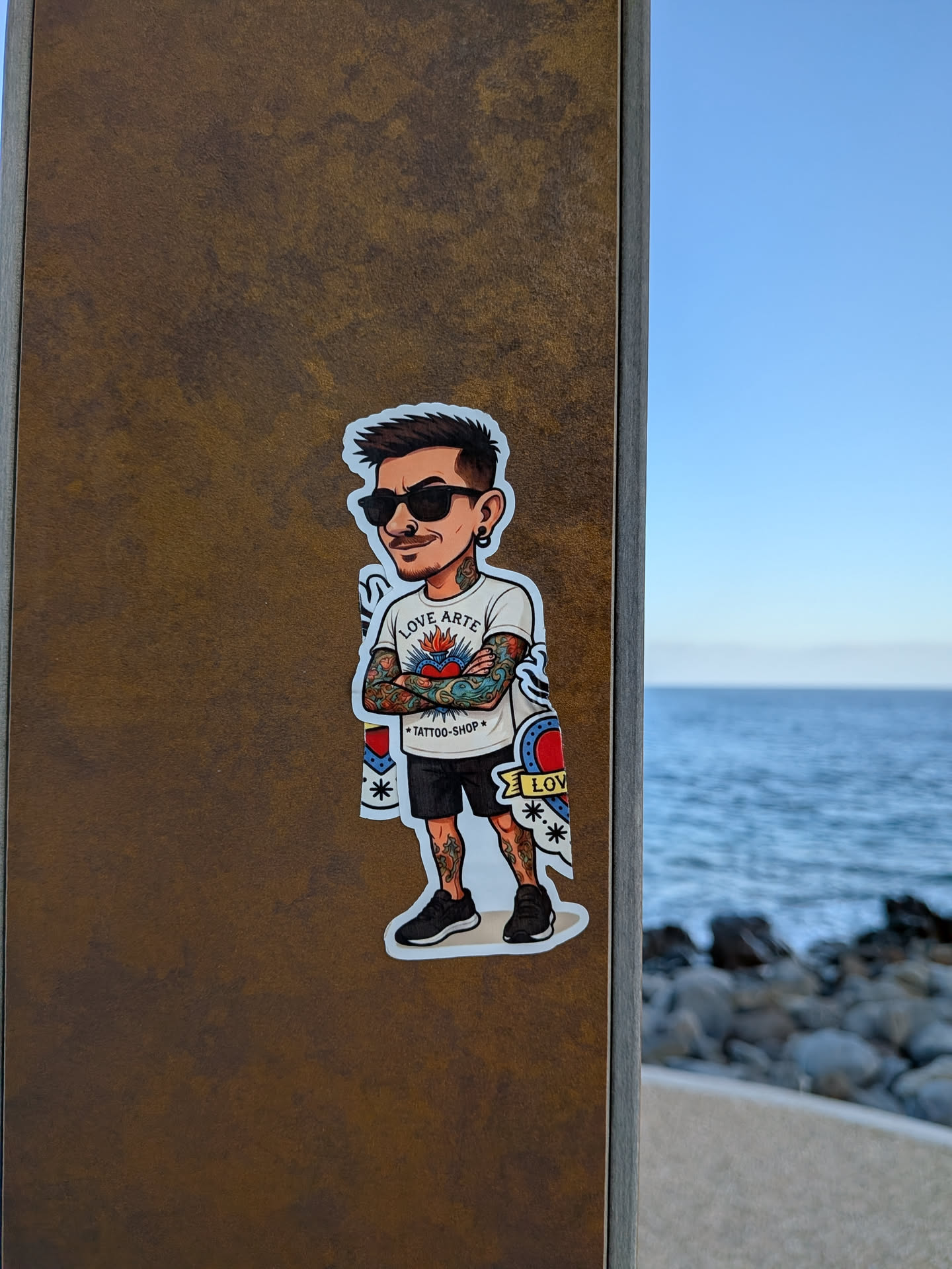 A die-cut sticker featuring a cartoon caricature of a man with spiky hair, sunglasses, and crossed arms. He is heavily tattooed on his arms and legs, wearing a white 'LOVE ARTE TATTOO-SHOP' t-shirt, black shorts, and black sneakers.