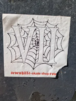 Street sticker A sticker featuring the Roman numerals VII (7) overlaid on a spiderweb design. The text 'sevenhills-skate shop ron' is visible at the bottom. The sticker appears to be placed on a dark gray surface with some texture.