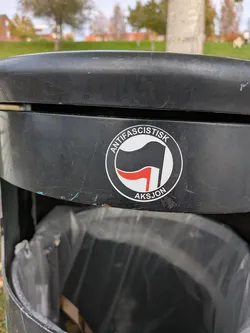 The sticker is circular with a white border. Inside, it features a black flag with a red stripe across it, held by a white pole. The text 'ANTIFASCISTISK' is written in black capital letters around the top curve of the circle, and 'AKSION' is written in black capital letters along the bottom curve. The background within the circle is white.