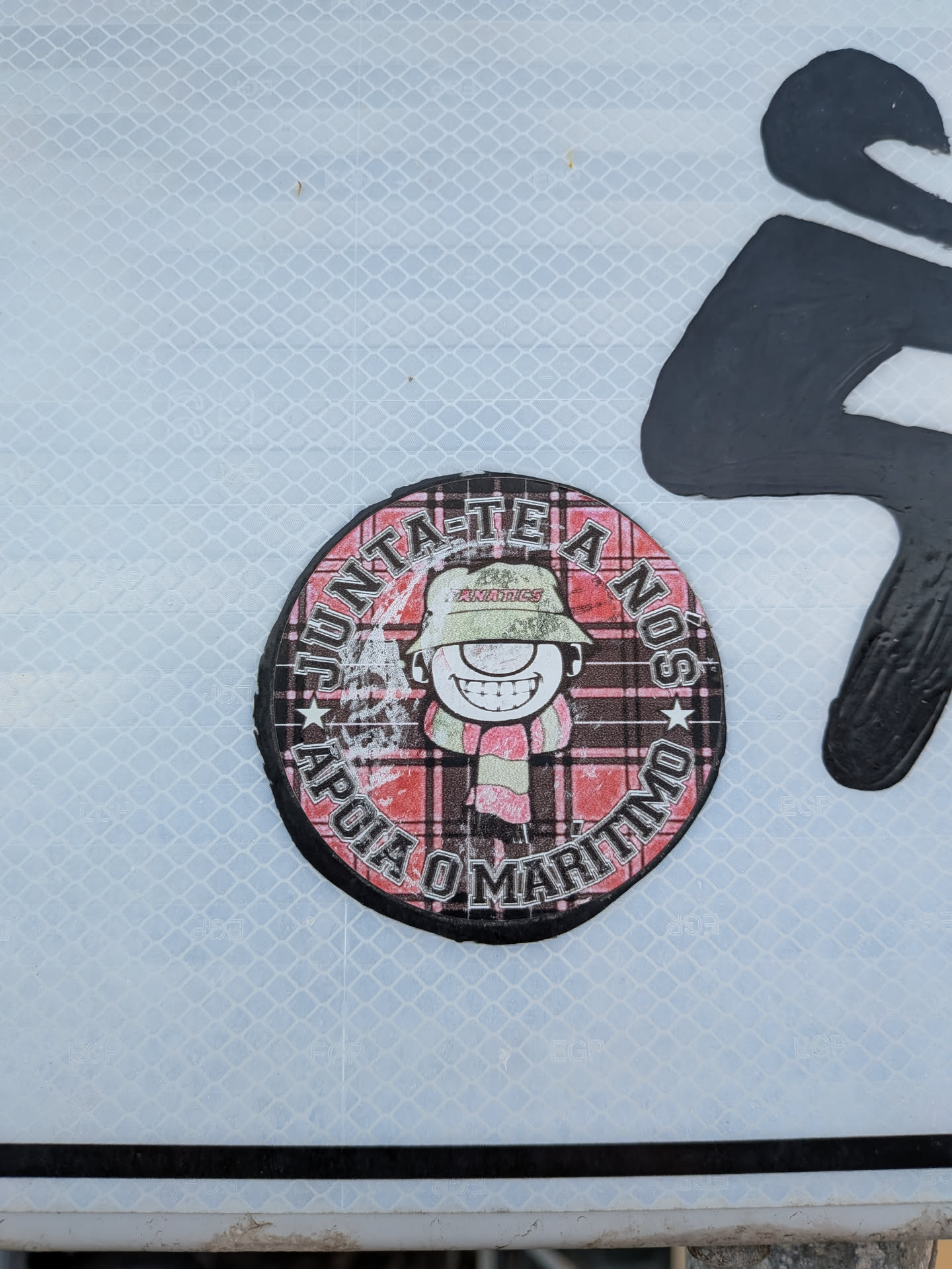 Circular sticker with a red plaid background. Features a smiling cartoon character wearing a green bucket hat and striped scarf, surrounded by Portuguese text: 'JUNTA-TE A NÓS' and 'APOIA O MARÍTIMO'.
