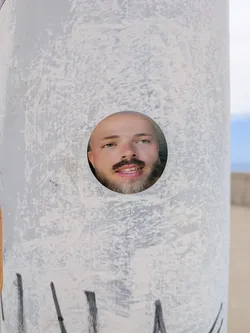 Street sticker A round sticker showing a photographic portrait of a man with a mustache and beard, looking off-camera. It is pasted on a textured, weathered pole.