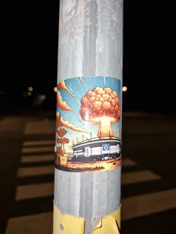 Street sticker A rectangular sticker affixed to a metal pole features a cartoon illustration. On the left, a brown mouse character resembling 'Die Maus' wearing a brown-and-white striped shirt presses down on a yellow TNT plunger detonator. On the right, the Volksparkstadion, bearing the blue and white diamond logo of Hamburger SV, is shown exploding with a massive orange and yellow mushroom cloud billowing into a blue sky.
