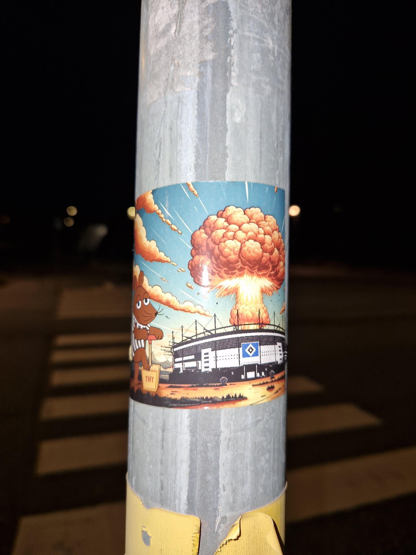 A rectangular sticker affixed to a metal pole features a cartoon illustration. On the left, a brown mouse character resembling 'Die Maus' wearing a brown-and-white striped shirt presses down on a yellow TNT plunger detonator. On the right, the Volksparkstadion, bearing the blue and white diamond logo of Hamburger SV, is shown exploding with a massive orange and yellow mushroom cloud billowing into a blue sky.
