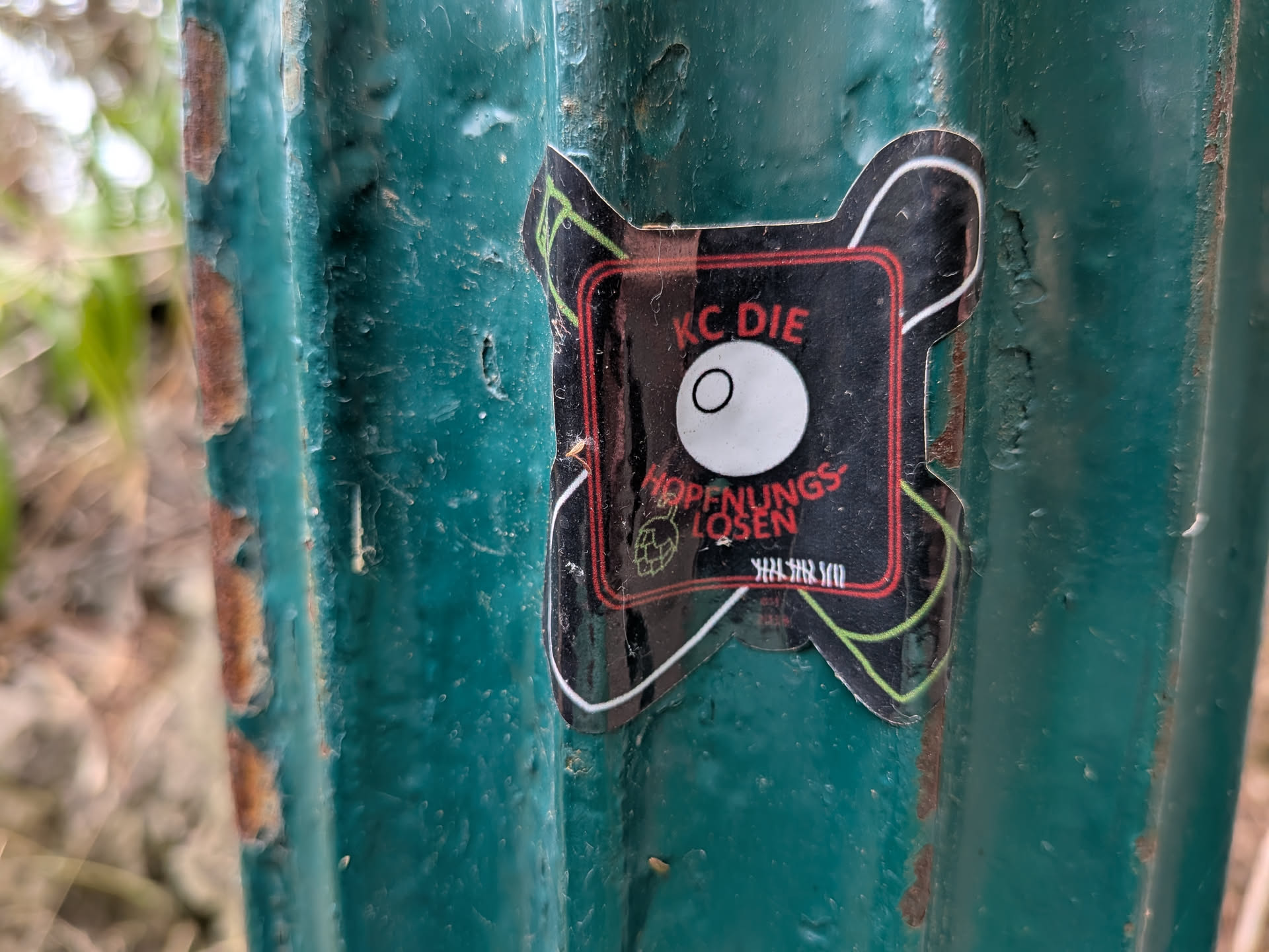 An X-shaped black sticker featuring a red square border. Inside, a central white eyeball graphic is flanked by a green hand grenade icon and white tally marks. The red text reads 'KC DIE HOFFNUNGSLOSEN'.