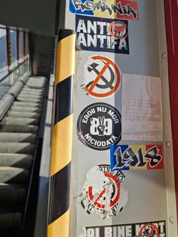 The image shows various stickers affixed to a metal pole, possibly in a public space.  The stickers depict various symbols and text, some appearing to be political or social commentary.  The central focus is a partially damaged sticker with illegible text and a symbol. Other stickers show anti-communist messages and a Romanian flag.