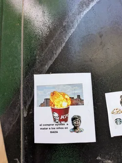 Street sticker A sticker featuring an altered KFC advertisement. The bucket of chicken has been replaced with an explosion over a building, likely meant to resemble bombings in Gaza. Below this imagery, there's text reading "al comprar ayudas a matar a los niños en GAZA," which translates to "buying helps kill children in GAZA." A crying child's face appears next to the bucket.