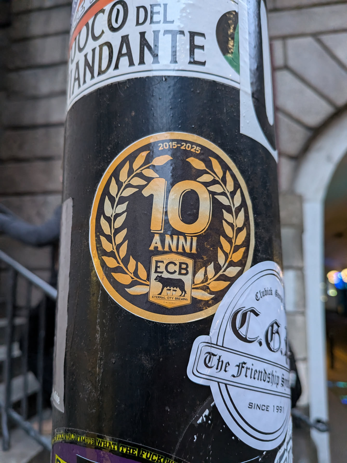 The sticker is circular with a prominent gold/yellow outer border and a black background. In the center, a large, stylized '10' is depicted in gold/yellow, encircled by a gold/yellow laurel wreath. Below the '10', the word 'ANNI' (Italian for years) is written in gold/yellow. Further below, a rectangular emblem features 'ECB' at the top, a silhouette of a she-wolf suckling two infants (a symbol of Rome) in the middle, and 'ETERNAL CITY BREWING' along with 'SINCE 2015' at the bottom, all in gold/yellow on a black background. Curved along the top edge, above the '10' and wreath, are the dates '2015-2025' in gold/yellow. The sticker shows some signs of wear, consistent with outdoor placement.