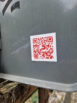 A red QR code is printed on a white square sticker, which is attached to a gray metal surface. The QR code has a small red symbol in the center.