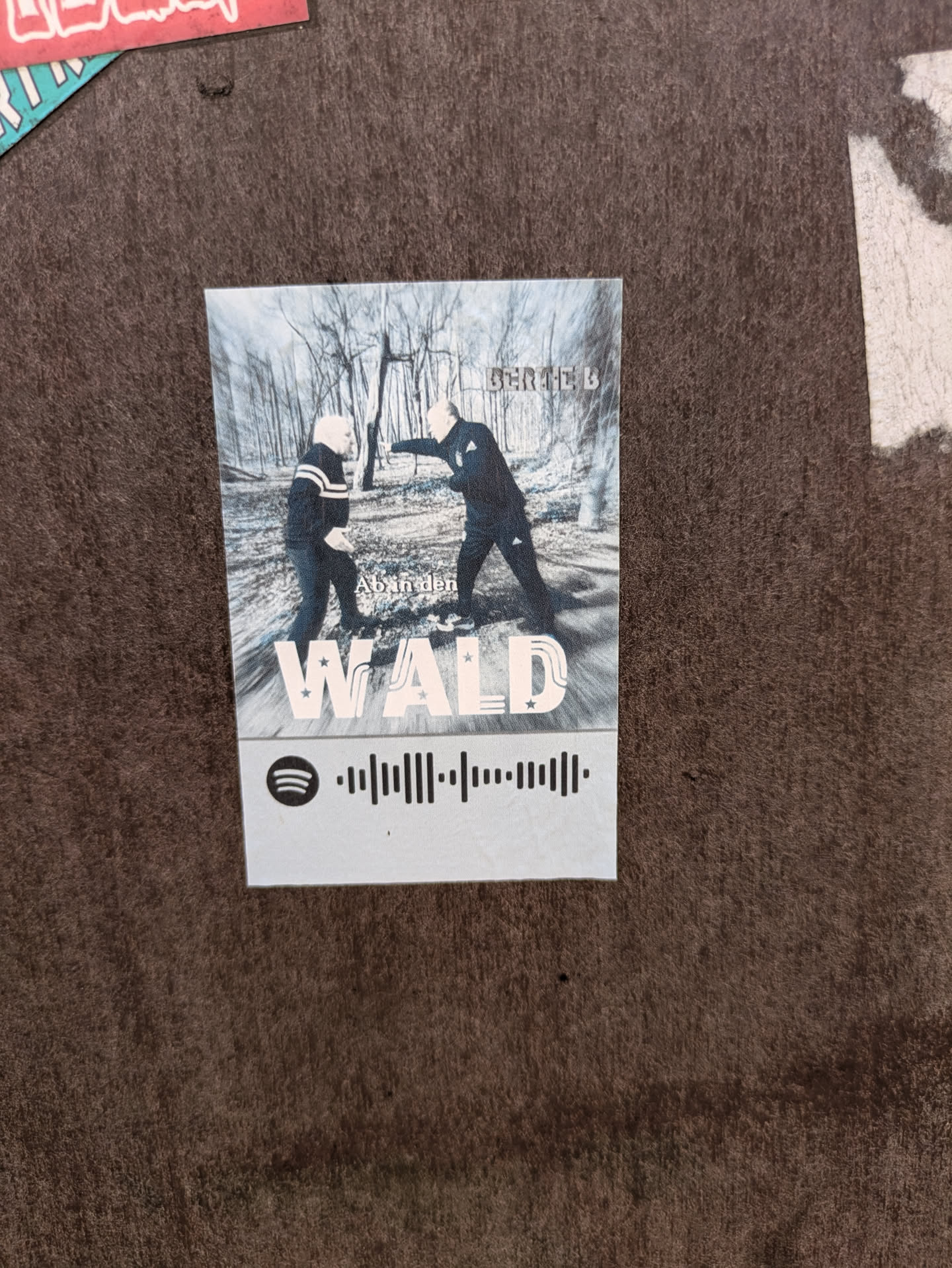 This sticker showcases what appears to be an album or single cover. The central image depicts two men dressed in dark attire, standing amidst a barren forest landscape, suggesting a winter or late autumn setting. One man on the right is gesturing or pointing towards the other. Prominently displayed is the text 'Ab in den WALD' (German for 'Off to the Forest' or 'Into the Forest') and 'BERTE B'. Below the image, a Spotify code is present, designed for direct scanning within the Spotify application to access the corresponding music track or album.