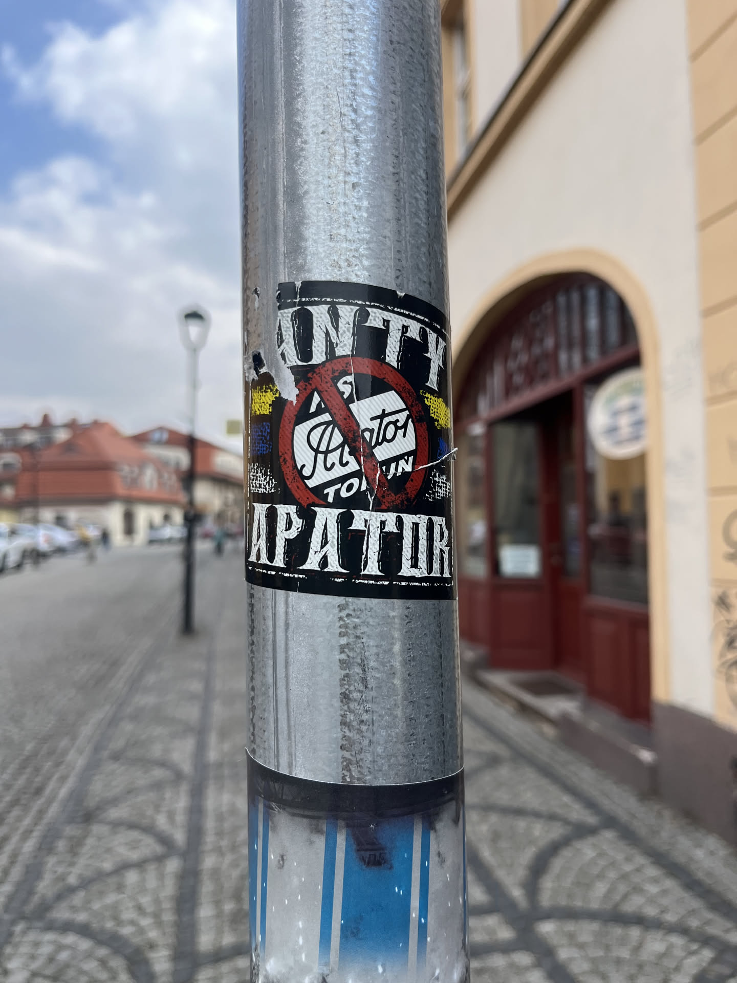 The sticker is rectangular with a distressed, grunge-like aesthetic, primarily in black and white with accents of red, yellow, and blue. At the top, partially obscured, is the word "UNTY" in bold, white block letters. The central feature is a large red circle with a diagonal line (the international 'no' symbol) overlaid on stylized white text that appears to read "Hator" and "TOUN". Below this central symbol, the word "APARTIR" is prominently displayed in large, distressed white block letters. To the left and right of the 'no' symbol, vertical stripes of yellow and blue are visible, reminiscent of the Ukrainian flag. The sticker shows signs of wear, with slight peeling at the edges and minor tears, indicating it is a physical sticker affixed to the metal pole.