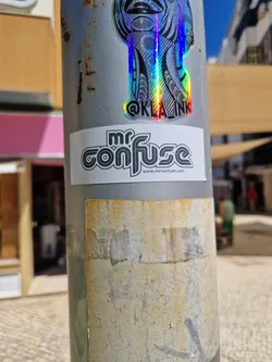 Street sticker A grayscale sticker with the text "Mr. Confuse" in a retro style font.  Below the main text, there's a smaller line of text indicating a website: www.mrconfuse.com. The sticker is attached to a pole, with other stickers visible nearby, including a holographic sticker with the Instagram handle @kla_ink.