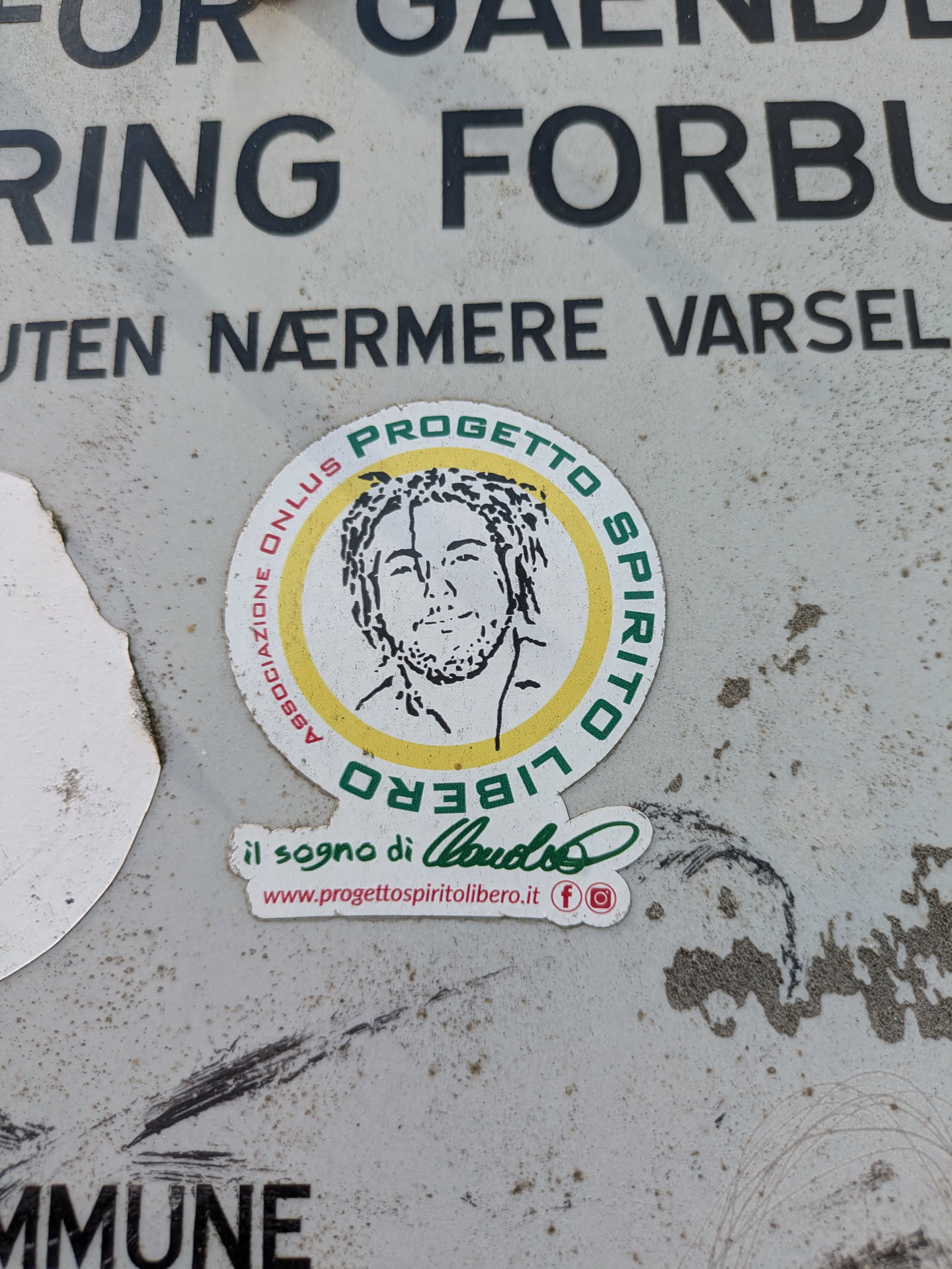 The sticker is primarily circular, featuring an outer green band with text 'PROGETTO SPIRITO LIBERO' (with 'LIBERO' inverted at the bottom) and 'ASSOCIAZIONE ONLUS' along the left side. Inside, a yellow ring frames a black-and-white line art illustration of a man with dreadlocks/curly hair and a beard. Directly below the main circle, a smaller, green-lettered text reads 'il sogno di Claudio' (the dream of Claudio), followed by the website 'www.progettospiritolibero.it' in red, and small red icons for Facebook and Instagram. The sticker is affixed to a textured, light grey surface that appears to be metal or a similar material, showing signs of dirt and wear.