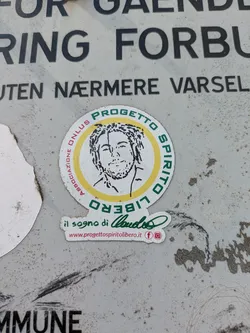 Street sticker The sticker is primarily circular, featuring an outer green band with text 'PROGETTO SPIRITO LIBERO' (with 'LIBERO' inverted at the bottom) and 'ASSOCIAZIONE ONLUS' along the left side. Inside, a yellow ring frames a black-and-white line art illustration of a man with dreadlocks/curly hair and a beard. Directly below the main circle, a smaller, green-lettered text reads 'il sogno di Claudio' (the dream of Claudio), followed by the website 'www.progettospiritolibero.it' in red, and small red icons for Facebook and Instagram. The sticker is affixed to a textured, light grey surface that appears to be metal or a similar material, showing signs of dirt and wear.