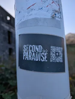 Street sticker A rectangular black sticker on a white post. It features the stylized white text 'SECOND PARADISE' intertwined with leafy vines, next to a QR code with a central star-like logo.