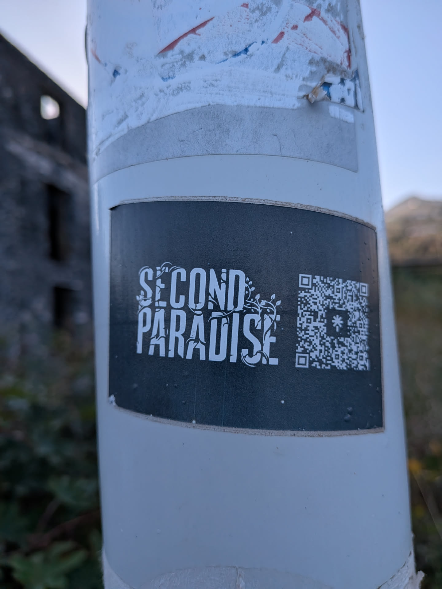 A rectangular black sticker on a white post. It features the stylized white text 'SECOND PARADISE' intertwined with leafy vines, next to a QR code with a central star-like logo.