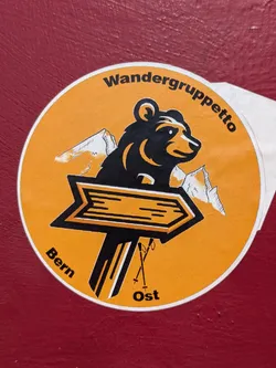 Street sticker The sticker is circular with a dominant orange background. It features a stylized black bear with some orange highlights, looking to the right. Behind the bear are two white, jagged mountain peaks, suggesting a snowy landscape. In front of the bear, at the bottom center, there is a black signpost shaped like an arrow pointing to the right, with orange accents. Leaning against the base of the signpost are two crossed ski poles. The text "Wandergruppetto" is curved along the top edge of the circle, while "Bern" is curved along the bottom left, and "Ost" is placed at the bottom right.