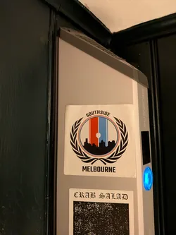 Street sticker The sticker is a white rectangle featuring a central circular emblem. This emblem is framed by a black laurel wreath. Inside the circle, the background is vertically striped in red (left), white (center), and light blue (right). A black silhouette of a city skyline, including a prominent tower, is depicted at the base of the circle. A small white heart shape is subtly placed within the white central stripe, above the skyline. Arched above the circular design, in black sans-serif font, is the word 'SOUTHSIDE'. Below the circle, in the same font, is 'MELBOURNE'. The sticker has a slightly worn appearance with minor creases.