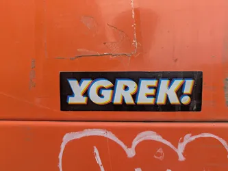Street sticker A sticker on an orange surface. The sticker has a black background with the word "YGREK!" in white with blue, yellow, and red 3D effects. There is some white graffiti below the sticker.