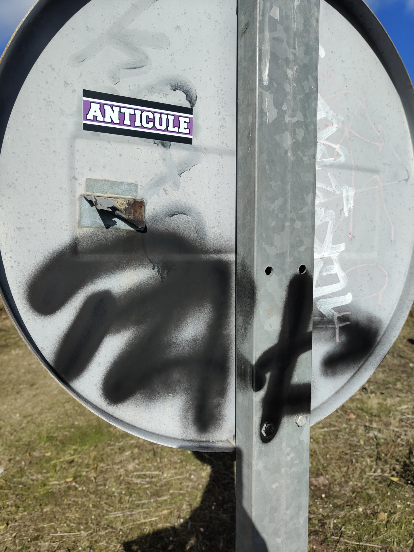 A rectangular sticker featuring the word "ANTICULE" in prominent, bold, white block letters with a thin black outline. The letters are set against a vibrant purple background, which is then framed by a thicker black border. The sticker is applied to a weathered, grey metal surface, which shows signs of wear, spray paint, and some rust.