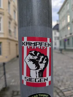 Street sticker A square-shaped sticker affixed to a gray metal pole. The design features a prominent black and white stylized raised fist, a common symbol of solidarity, power, and resistance. Above the fist, in bold, blocky capital letters, is the German phrase "KÄMPFEN UND" (Fight and) and below the fist, "SIEGEN" (Win/Conquer). The background of the sticker is composed of red and white stripes, forming a cross pattern similar to a Nordic flag (like the Danish flag), with a white cross on a red field and white borders on the top and bottom. The overall aesthetic is one of determination and defiance.