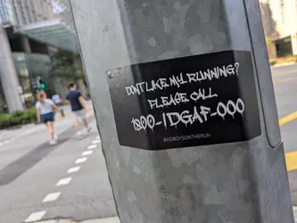 Street sticker Black rectangular sticker on a metal pole. Features white dripping graffiti text reading 'DON'T LIKE MY RUNNING? PLEASE CALL 1800-IDGAF-000' and 'BADBOYSONTHERUN' in small print below.