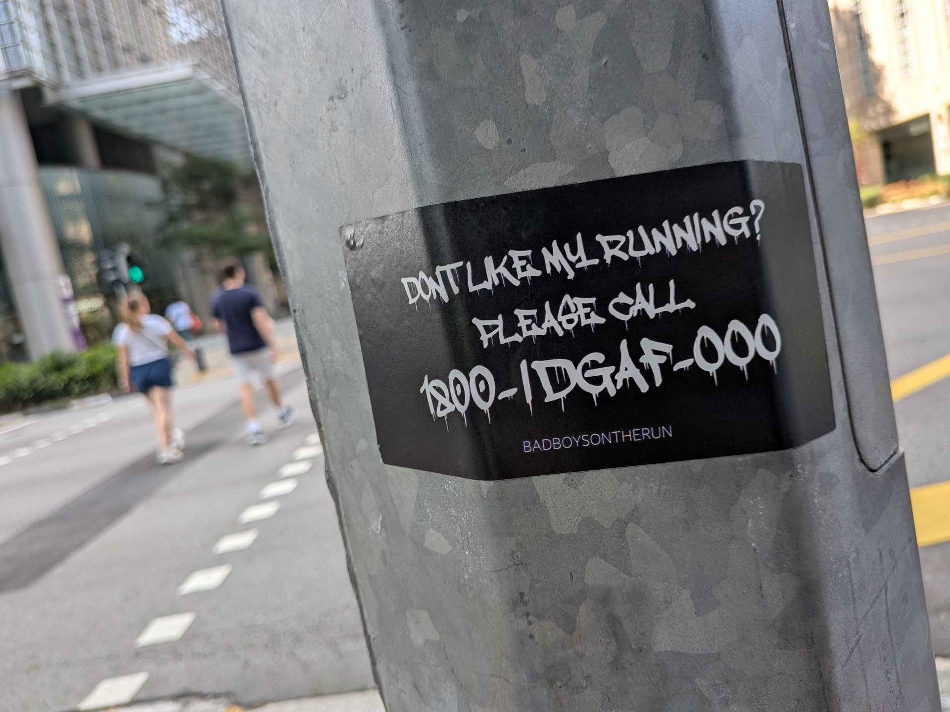 Black rectangular sticker on a metal pole. Features white dripping graffiti text reading 'DON'T LIKE MY RUNNING? PLEASE CALL 1800-IDGAF-000' and 'BADBOYSONTHERUN' in small print below.