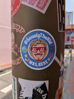 Street sticker A round blue sticker with a central cartoon portrait of a smiling man wearing gradient sunglasses and sporting curly blonde hair. Surrounding the portrait are small illustrations including a globe, a map outline, a fox, a watermelon slice, and a QR code. Text reads '@SoualyTheKid WAS HERE' in white.
